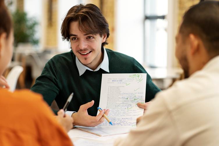 Smiling young man holding a graded paper with handwritten notes and check marks while discussing feedback with two people across a table in a bright indoor setting.