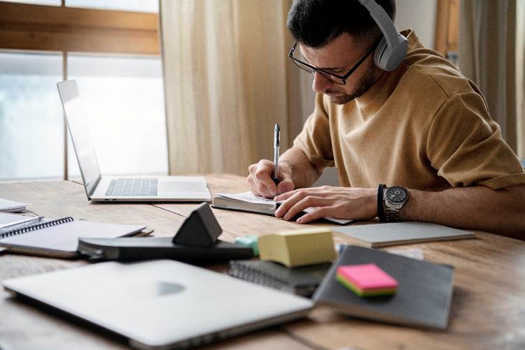 A focused man wearing headphones working on a laptop and writing notes at a wooden desk, representing productivity, online learning, and remote work.