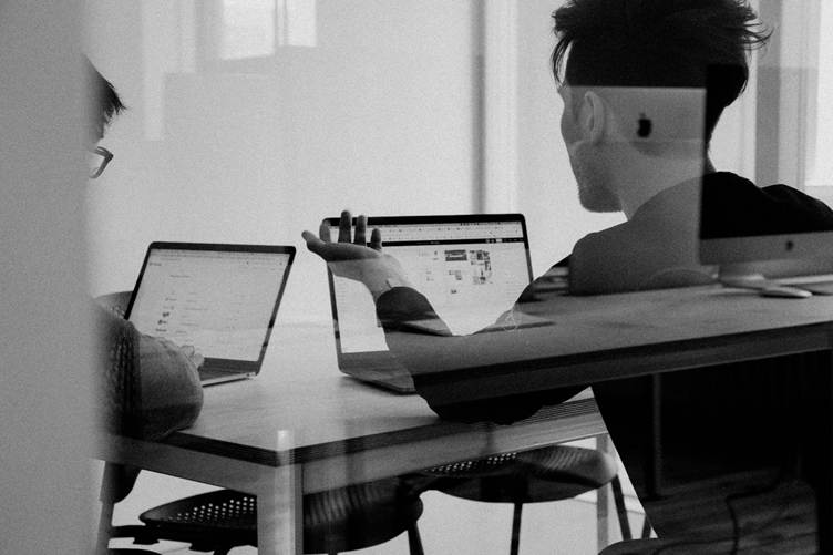 Two people seated at a table working on laptops, engaged in a discussion inside a modern office or study space, viewed through a glass partition in black and white.