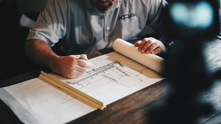 A professional designer sketches a detailed architectural plan on paper using a pencil and ruler, working at a wooden desk under focused lighting.