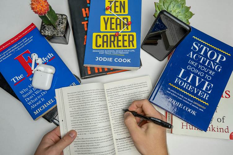 A flat-lay of career and self-development books on a desk, including “Ten Year Career” and “Stop Acting Like You’re Going to Live Forever,” with a person taking notes in an open book, surrounded by a phone, earbuds, and small plants.
