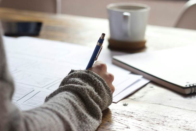 Person writing notes on paper at a wooden desk with a pen, notebook, and coffee cup in a cozy workspace setting.