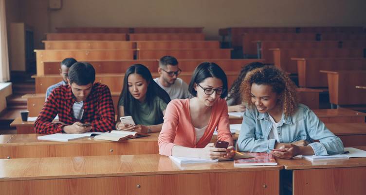 Group of college students sitting in a lecture hall, using smartphones and studying together at wooden desks with notebooks and textbooks.
