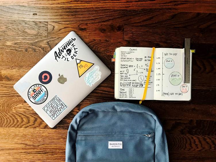 Alt text:
Top view of a laptop with stickers, an open notebook filled with study notes and diagrams, and a blue backpack arranged on a wooden floor.