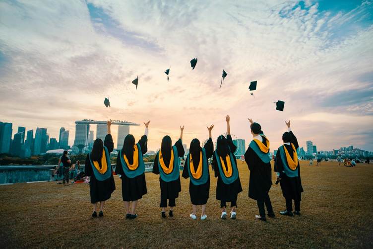 A group of graduates wearing black gowns with yellow stoles stand on a grassy field at sunset, tossing their graduation caps into the air, with a modern city skyline in the background.