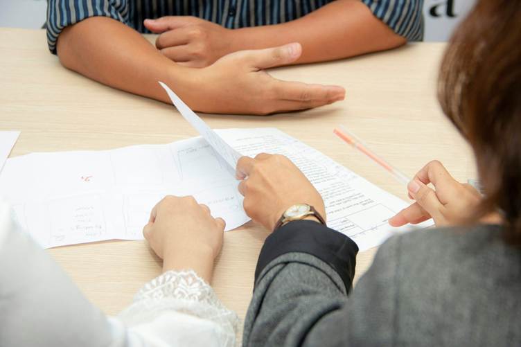 Two people sitting at a desk reviewing documents, with one person pointing at a paper while holding a pen during a discussion or interview.