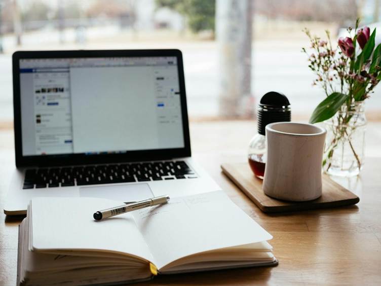 A laptop open on a wooden desk beside an open notebook with a pen, a white coffee mug on a tray, and a small vase of flowers near a window, creating a cozy workspace setting.