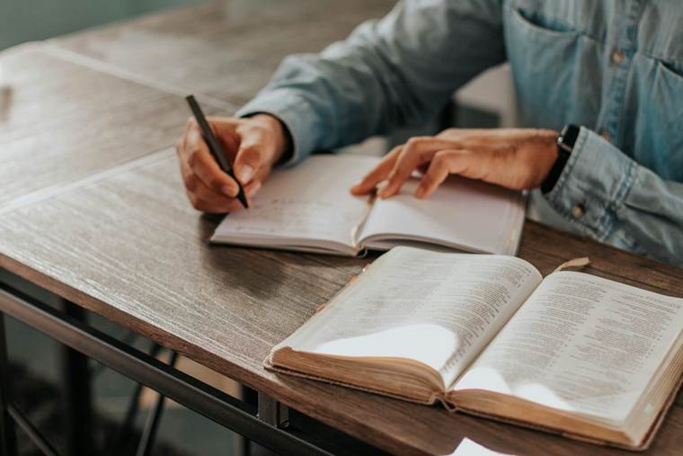 Person wearing a denim shirt writing in a notebook at a wooden table, with an open book placed in front for reference.