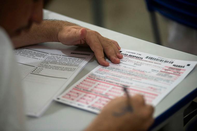 Alt text: Close-up of a person filling out a multiple-choice answer sheet with a pen during an exam, with one hand resting on the desk and the test paper visible.