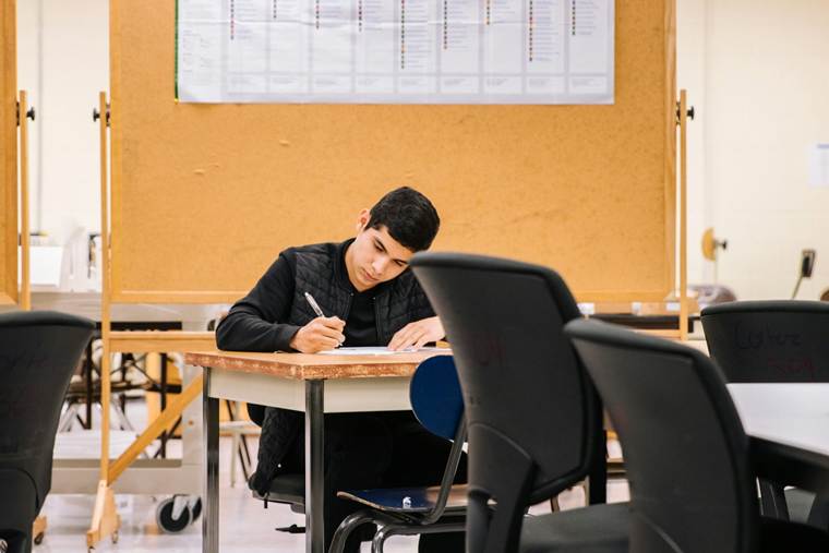 Student sitting alone at a classroom desk, writing on paper with focused concentration, with empty chairs and a notice board in the background.