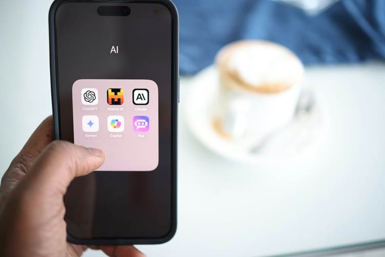 A hand holding a smartphone displaying a folder labeled “AI” with apps like ChatGPT, Mistral AI, Claude, Gemini, Copilot, and Poe on the screen, with a blurred coffee cup in the background.
