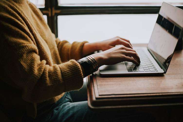 Person wearing a brown sweater typing on a laptop at a wooden desk near a window, with natural light coming in from the side.