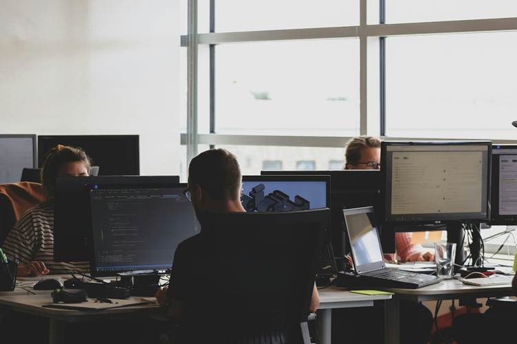 Team of people working at desktop computers in a modern office, with large monitors displaying code and documents near a bright window.