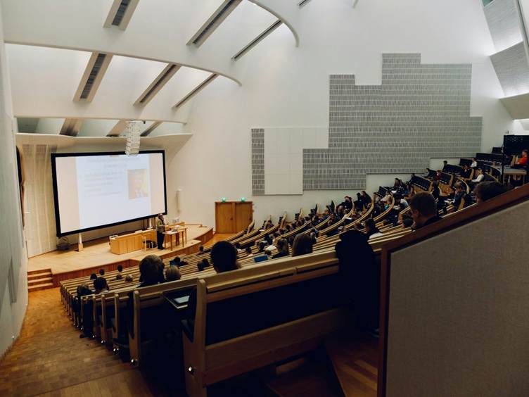 A large modern university lecture hall with tiered wooden seating, students seated and facing a big projection screen at the front where a presenter stands at a podium. The room has high white ceilings with skylights and acoustic panels on the walls.