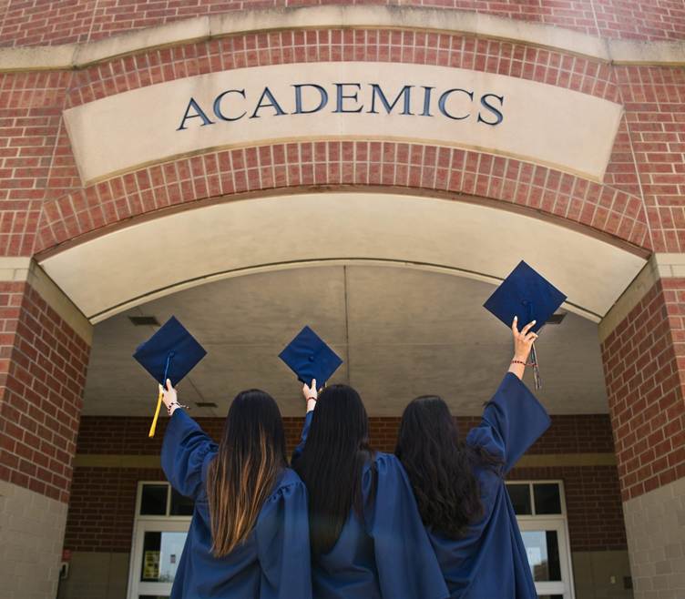 Three graduates in blue caps and gowns stand with their backs to the camera, raising their mortarboards in celebration beneath a brick archway labeled “ACADEMICS” at a college building entrance.