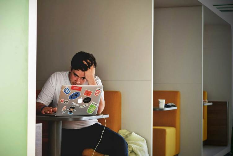 A stressed man sits alone in a small booth workspace, holding his head while working on a laptop covered with programming stickers like PHP, GitHub, HTML5, and Node.js, with a coffee cup on a nearby table.
