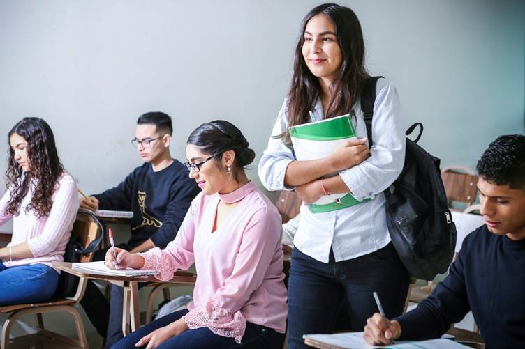 A group of college students in a classroom, with three students seated and writing in notebooks while a smiling female student stands holding a green notebook and wearing a backpack.