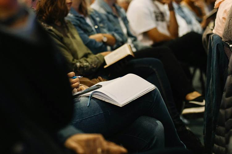 Students seated in a classroom or seminar setting, holding notebooks and books, with one person writing notes on a notepad while listening attentively.