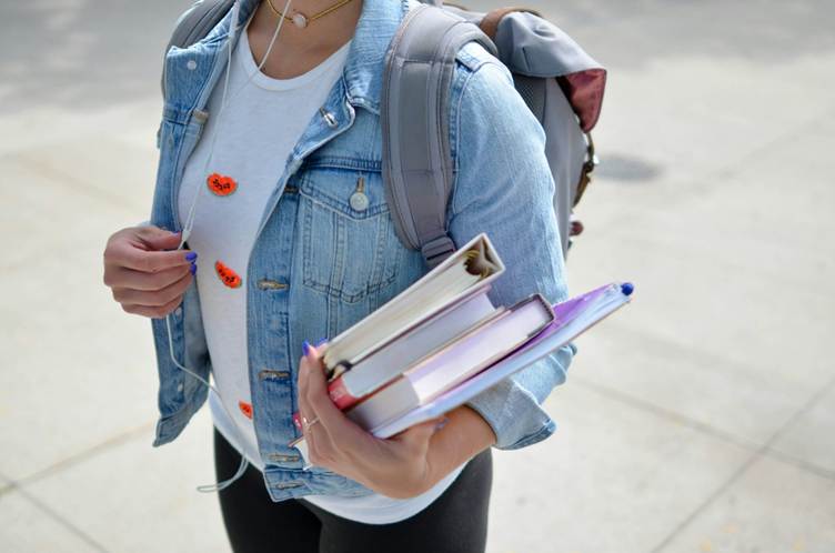 College student wearing a denim jacket and backpack, holding textbooks and a notebook while listening to earphones outdoors on campus.