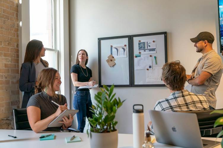 Team of five professionals collaborating in a modern office, with a woman presenting ideas on a wall board while others take notes and listen around a table with laptops and a plant.