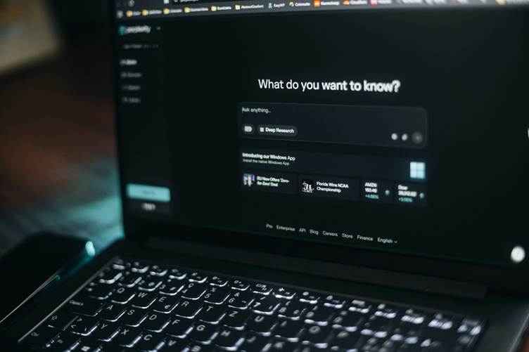 Close-up of a laptop displaying an AI chatbot interface with the prompt “What do you want to know?” and a search bar, viewed in a dimly lit setting with the keyboard illuminated.