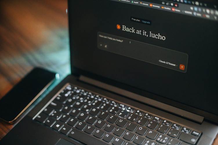 Laptop on a wooden desk displaying an AI assistant interface with the greeting “Back at it” and a text input field, with the keyboard illuminated and a smartphone placed beside it.