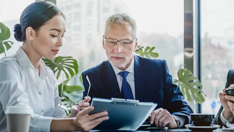 “Senior business professional reviewing documents on a tablet with a colleague during a professional office meeting, discussing strategy and data analysis.”