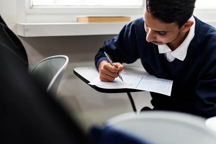 A student wearing a dark blue sweater sits at a small classroom desk, writing on a test paper with a pen. The student is focused on the exam sheet, while other desks and a blurred classmate appear in the foreground. Natural light comes from a window behind the desk, creating a quiet exam-room atmosphere. ✏️📄