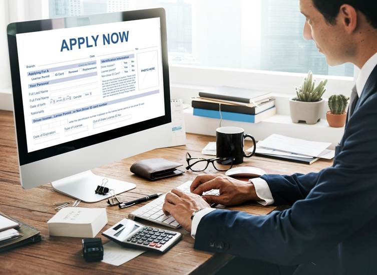 Professional man in a suit sitting at a desk and typing on a computer, with an “Apply Now” online application form displayed on the screen, alongside office items like a calculator, notebook, coffee mug, and glasses.