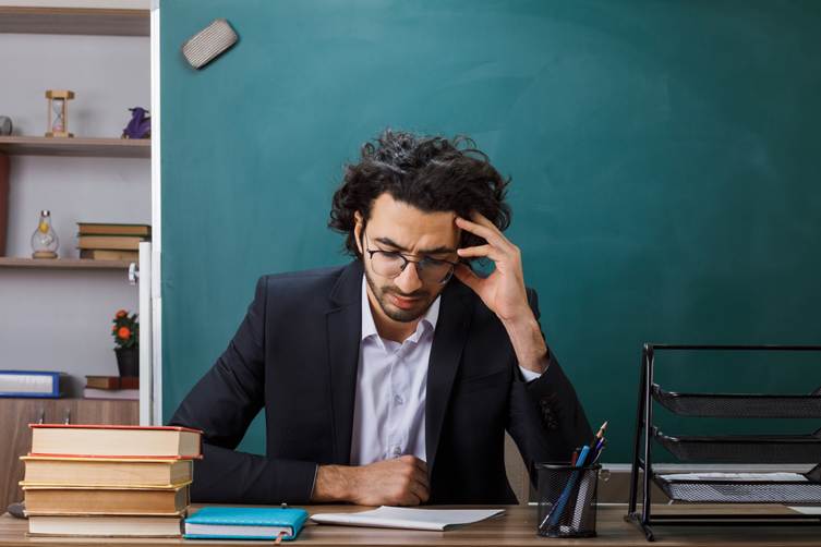 “Student sitting at a desk with books, holding his head while studying, appearing stressed or deep in concentration in front of a classroom chalkboard.”