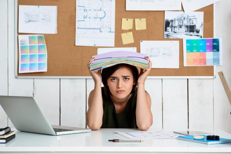 A stressed student sitting at a desk holds a stack of books on their head while looking frustrated. A laptop and study materials are on the table, and a corkboard with notes, sketches, and color charts is displayed on the wall behind them, suggesting academic pressure and heavy study workload.
