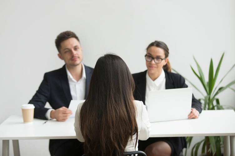 A job candidate with long dark hair sits facing two interviewers in a professional office setting, while the interviewers review documents and a laptop during the interview.